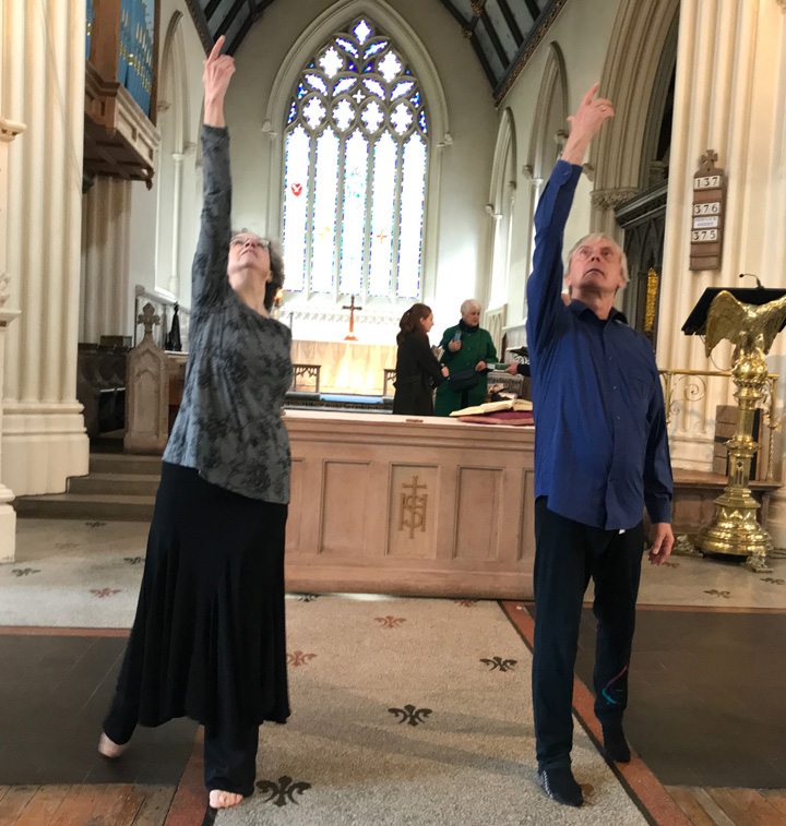 Two dancers have right arms raised and are looking upwards. They are in front of an altar and an east window