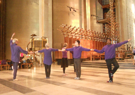 six standing dancers, dressed in purple and black, make a large wheel figure by linking arms
